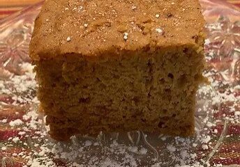 A square slice of brown cake dusted with powdered sugar on a glass plate.