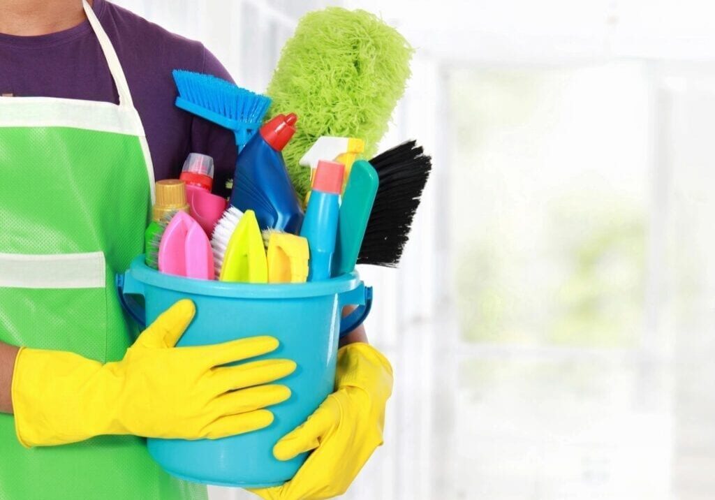 Person holding a bucket filled with colorful cleaning supplies and tools.