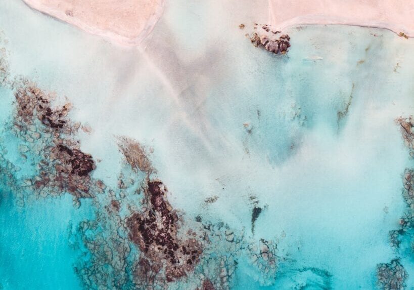 Aerial view of turquoise ocean meeting sandy pink beach with coral patches.
